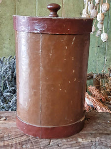 Antique German apothecary jar (brown with maroon stripes) on a rustic stool. It is styled with a black bucket of lavender and a vineyard basket against a sage green planked door.