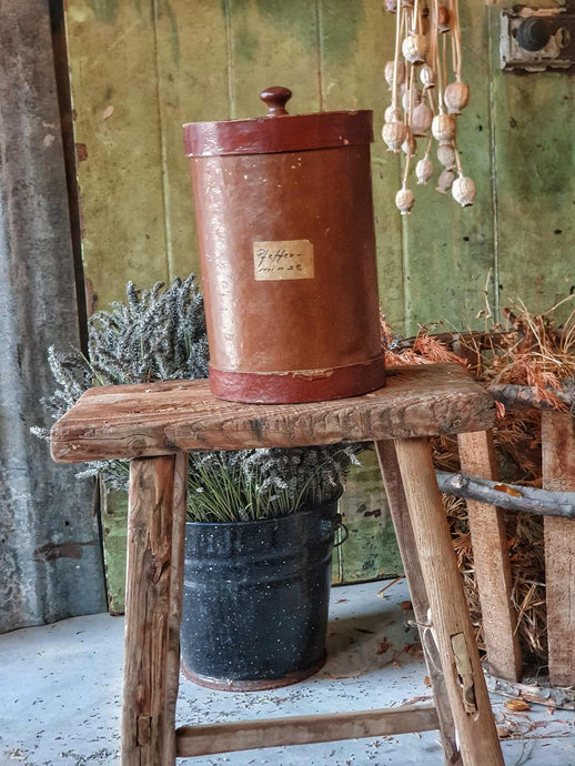 Antique German apothecary jar (brown with maroon stripes) on a rustic stool. It is styled with a black bucket of lavender and a vineyard basket against a sage green planked door.
