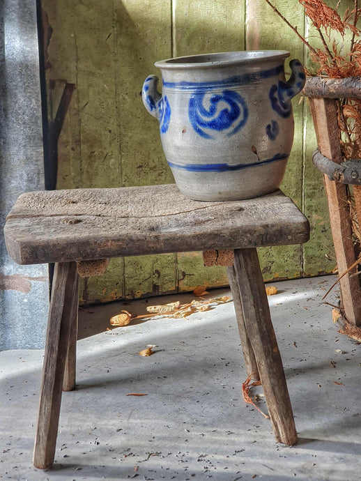 Grey and blue Betschdorf confit pot sitting on a low, weathered rustic farmhouse stool. In the background is a rustic farmhouse door painted in sage green, with sunlight streaming in from the left.
