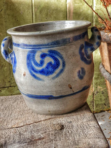 Grey and blue Betschdorf confit pot sitting on a low, weathered rustic farmhouse stool. In the background is a rustic farmhouse door painted in sage green, with sunlight streaming in from the left.