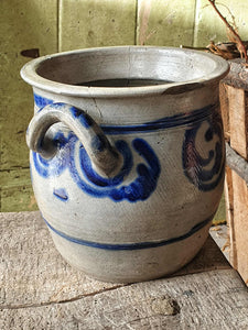 Grey and blue Betschdorf confit pot sitting on a low, weathered rustic farmhouse stool. In the background is a rustic farmhouse door painted in sage green, with sunlight streaming in from the left.