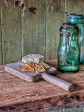 Load image into Gallery viewer, Antique Hungarian oak chopping board with a distinctive tulip-shaped outline and long handle, displayed with cheese and rustic food on a weathered wooden table. The solid oak board has a few historic cutting scars and natural aged wood texture. A green glass jar and rustic Hungarian and European farmhouse kitchen styling sit in the background, creating a warm French country and folk-art inspired atmosphere. The board measures 13 inches (33 cm) long including the handle and 6.5 inches (16.5 cm) wide.