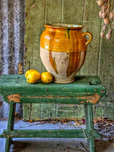 Load image into Gallery viewer, A rare early 19th-century French confit pot with a single arched handle and a footed base. The antique terracotta crock features a rich caramel-mustard and honey-colored glaze with dramatic vertical kiln runs and pooling at the rim, styled on an old painted stool against weathered green boards with fresh lemons.
