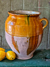 Load image into Gallery viewer, A rare early 19th-century French confit pot with a single arched handle and a footed base. The antique terracotta crock features a rich caramel-mustard and honey-colored glaze with dramatic vertical kiln runs and pooling at the rim, styled on an old painted stool against weathered green boards with fresh lemons.