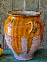 Load image into Gallery viewer, A rare early 19th-century French confit pot with a single arched handle and a footed base. The antique terracotta crock features a rich caramel-mustard and honey-colored glaze with dramatic vertical kiln runs and pooling at the rim, styled on an old painted stool against weathered green boards with fresh lemons.