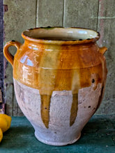 Load image into Gallery viewer, A rare early 19th-century French confit pot with a single arched handle and a footed base. The antique terracotta crock features a rich caramel-mustard and honey-colored glaze with dramatic vertical kiln runs and pooling at the rim, styled on an old painted stool against weathered green boards with fresh lemons.