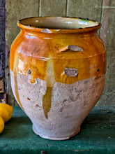 Load image into Gallery viewer, A rare early 19th-century French confit pot with a single arched handle and a footed base. The antique terracotta crock features a rich caramel-mustard and honey-colored glaze with dramatic vertical kiln runs and pooling at the rim, styled on an old painted stool against weathered green boards with fresh lemons.