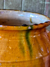 Load image into Gallery viewer, A rare early 19th-century French confit pot with a single arched handle and a footed base. The antique terracotta crock features a rich caramel-mustard and honey-colored glaze with dramatic vertical kiln runs and pooling at the rim, styled on an old painted stool against weathered green boards with fresh lemons.