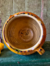 Load image into Gallery viewer, A rare early 19th-century French confit pot with a single arched handle and a footed base. The antique terracotta crock features a rich caramel-mustard and honey-colored glaze with dramatic vertical kiln runs and pooling at the rim, styled on an old painted stool against weathered green boards with fresh lemons.