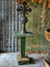 Load image into Gallery viewer, An ornate dark metal crucifix with a ribbed stem and decorative cross, mounted on a weathered green pedestal table against a rustic corrugated metal and wood wall backdrop.