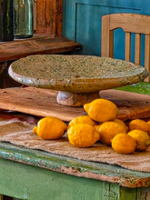 Load image into Gallery viewer, A heavy, hand-formed Moroccan Tamegroute footed serving dish with a soft cream ground and a delicate green splatter glaze. The shallow pedestal bowl features a rough, textured, salt-fired crystalline finish and is styled filled with bright lemons against a backdrop of weathered timber and rustic country-house furniture.