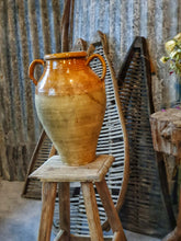 Load image into Gallery viewer, A 15-inch 19th-century Southern French olive jar with a glowing, deep mustard glaze. The jar shows an honest patina of chips and historical repairs. It is styled in a lifestyle setting in front of weathered wooden fruit drying trays, highlighting its use as a rustic home decor centerpiece.