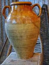 Load image into Gallery viewer, A 15-inch 19th-century Southern French olive jar with a glowing, deep mustard glaze. The jar shows an honest patina of chips and historical repairs. It is styled in a lifestyle setting in front of weathered wooden fruit drying trays, highlighting its use as a rustic home decor centerpiece.