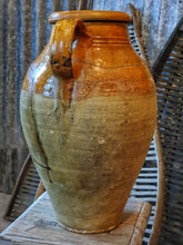 Load image into Gallery viewer, A 15-inch 19th-century Southern French olive jar with a glowing, deep mustard glaze. The jar shows an honest patina of chips and historical repairs. It is styled in a lifestyle setting in front of weathered wooden fruit drying trays, highlighting its use as a rustic home decor centerpiece.