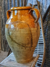 Load image into Gallery viewer, A 15-inch 19th-century Southern French olive jar with a glowing, deep mustard glaze. The jar shows an honest patina of chips and historical repairs. It is styled in a lifestyle setting in front of weathered wooden fruit drying trays, highlighting its use as a rustic home decor centerpiece.