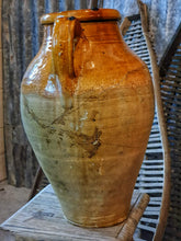 Load image into Gallery viewer, A 15-inch 19th-century Southern French olive jar with a glowing, deep mustard glaze. The jar shows an honest patina of chips and historical repairs. It is styled in a lifestyle setting in front of weathered wooden fruit drying trays, highlighting its use as a rustic home decor centerpiece.
