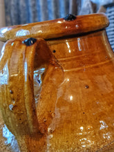 Load image into Gallery viewer, A 15-inch 19th-century Southern French olive jar with a glowing, deep mustard glaze. The jar shows an honest patina of chips and historical repairs. It is styled in a lifestyle setting in front of weathered wooden fruit drying trays, highlighting its use as a rustic home decor centerpiece.